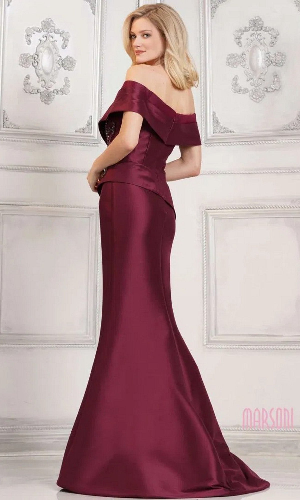 A woman in the Marsoni MV1312 long formal dress—a stretch Mikado, off-shoulder, floor-length gown with a mermaid skirt—stands in a white, ornately decorated room, facing slightly away from the camera.
